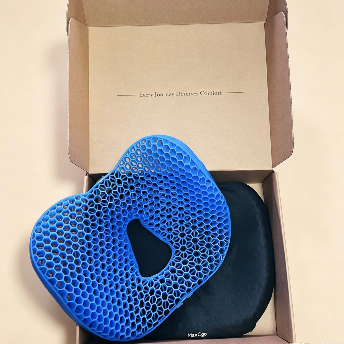 A blue honeycomb mesh ergonomic cushion with a central cutout, displayed inside an open brown cardboard box next to a black fabric pouch. The box interior is printed with the text 'Every Journey Deserves Comfort'.