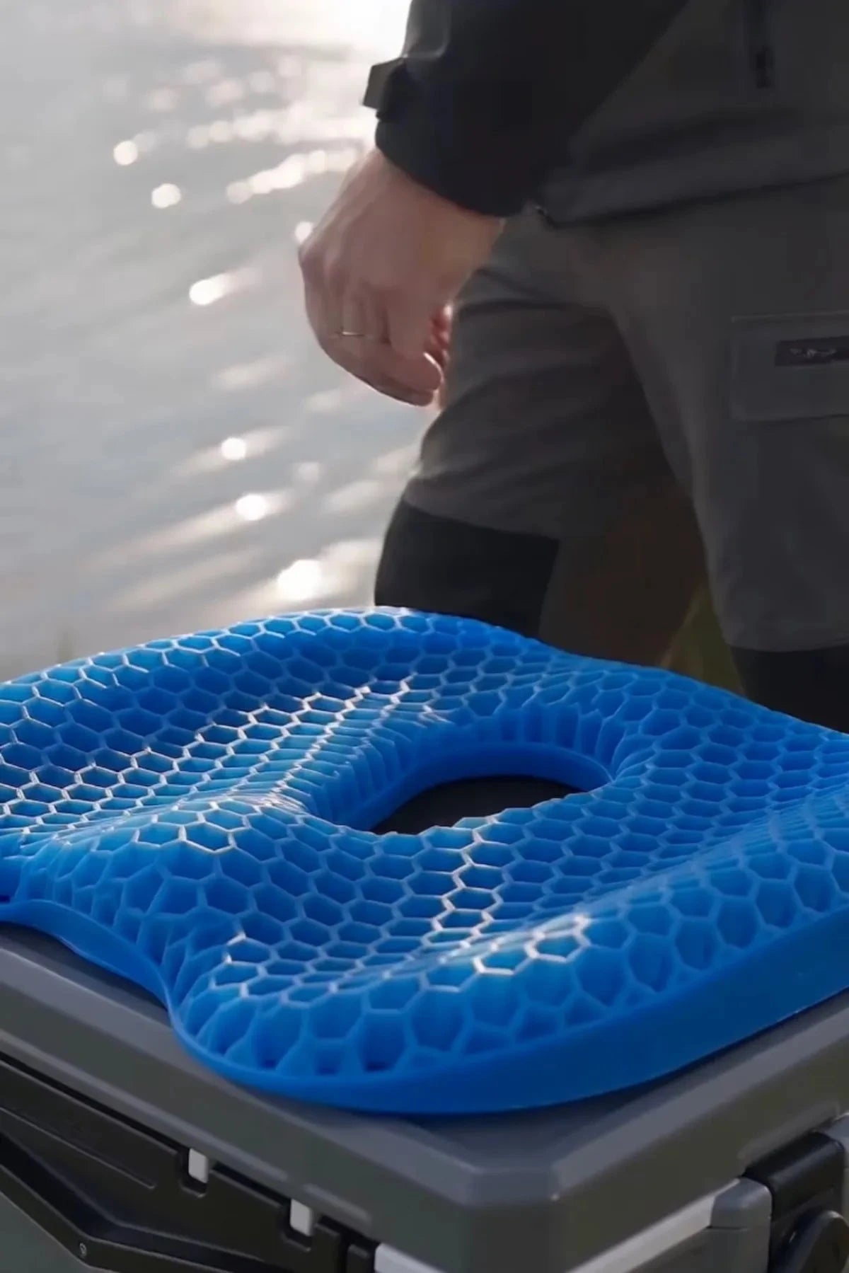 A bright blue contoured seat pad with a honeycomb texture resting on top of a gray cooler, with a person standing in the background near water.