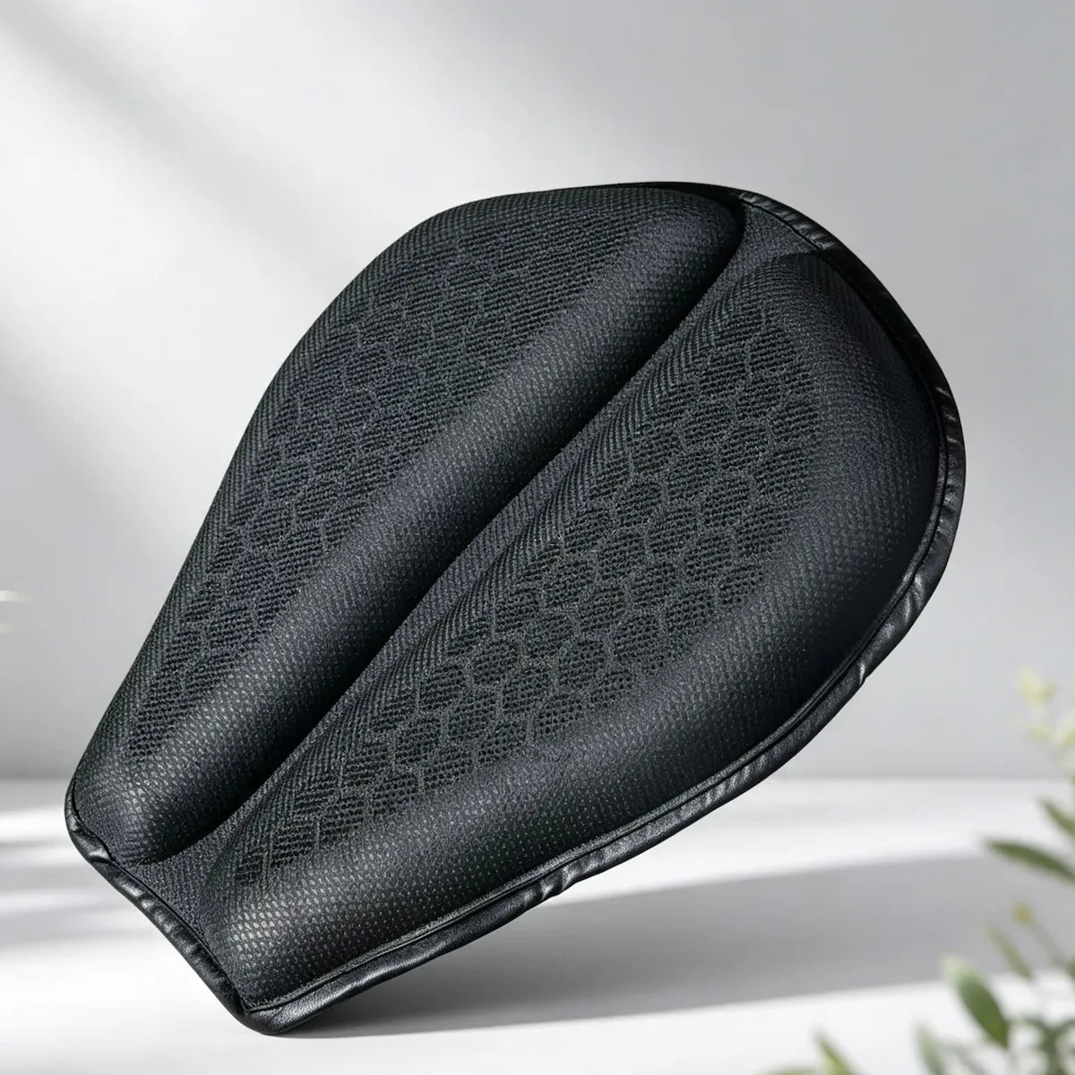 A black motorcycle or bicycle handlebar grip cover with a textured honeycomb pattern, shown floating against a light gray background with soft shadows and blurred green leaves in the corner.