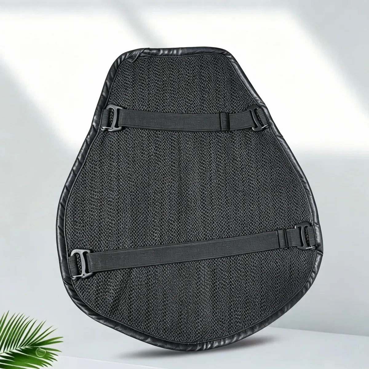 A black motorcycle or scooter seat cushion with a textured mesh center and two adjustable black straps, shown on a light gray background with a small green plant leaf in the corner.