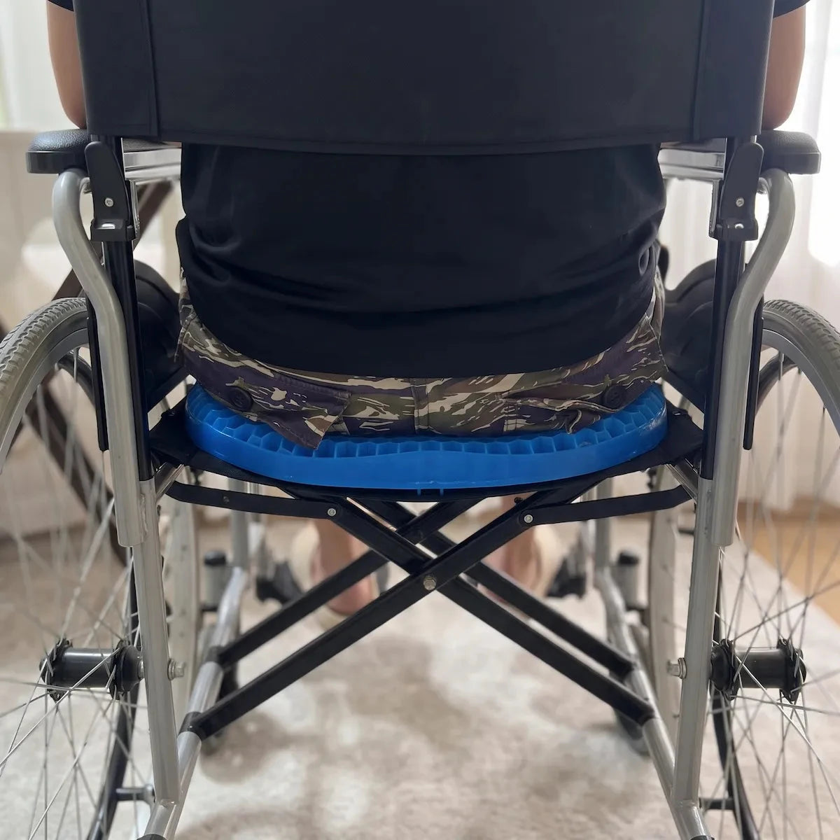A person seated in a wheelchair viewed from behind, wearing a bright blue contoured seat pad with camouflage-patterned fabric around the waist, in an indoor setting.