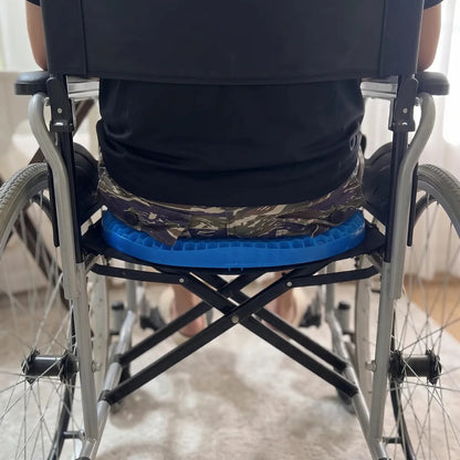 A person seated in a wheelchair viewed from behind, wearing a bright blue contoured seat pad with camouflage-patterned fabric around the waist, in an indoor setting.